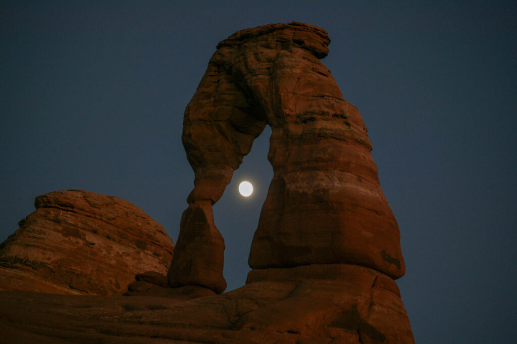 Moon framed by Delicate Arch at night.